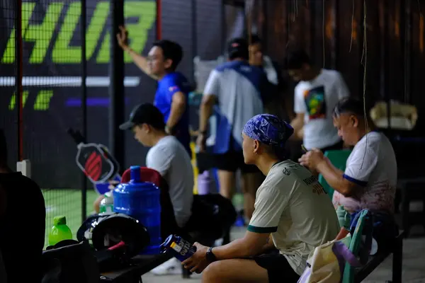 A group of players resting courtside on benches at RC Rally Hub, one wearing a blue bandana, with paddles and water jugs nearby