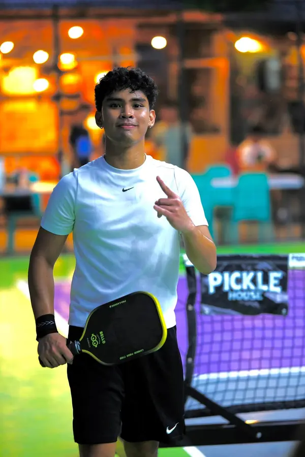A young man in a white Nike shirt smiling and pointing upward, holding a paddle, with a Pickle House court sign behind him