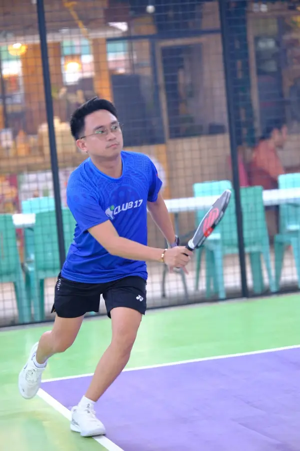 A man in a blue Club 17 jersey and glasses making a backhand return on the court