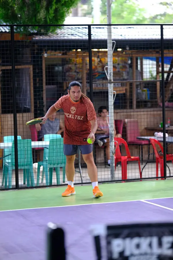 A man in an orange Pickltgz jersey preparing an underhand serve on the court at RC Rally Hub