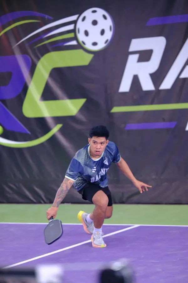 A man in a patterned team jersey reaching low for a return shot, RC Rally Hub banner behind him