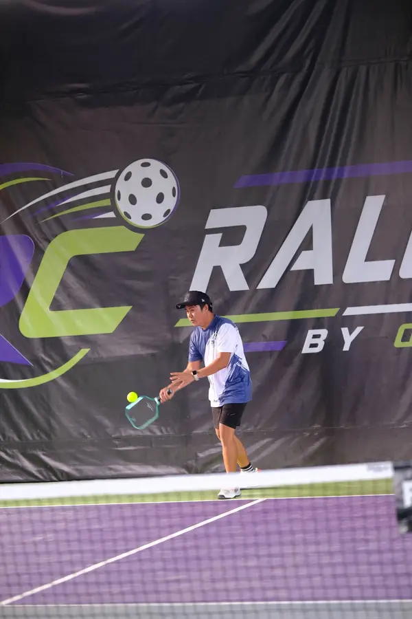 A man in a blue and white jersey and black cap making a forehand return near the net, RC Rally Hub banner behind him