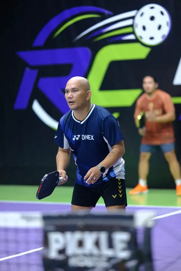 A bald man in a navy Yonex jersey holding a paddle ready at the net, RC Rally Hub step-and-repeat banner behind him, an opponent in orange blurred in the background